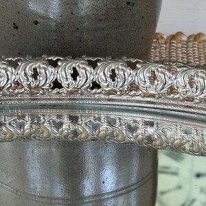 May include: A silver-toned oval tray featuring a raised, intricate border with a repeating embossed scroll and leaf pattern. The metallic rim has a polished finish, and the central area is reflective, showing a clear mirror-like surface.