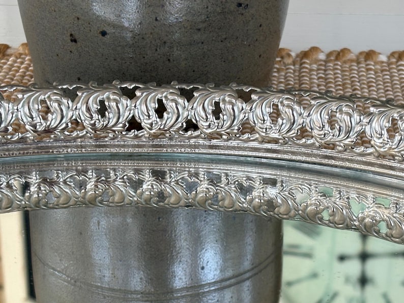 May include: A silver-toned oval tray featuring a raised, intricate border with a repeating embossed scroll and leaf pattern. The metallic rim has a polished finish, and the central area is reflective, showing a clear mirror-like surface.