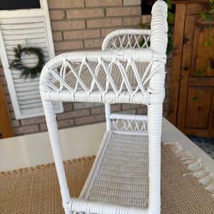 May include: A white wicker shelf with a rectangular base and a curved top. The shelf features a woven design and is supported by vertical posts. The piece is designed for storage or display and has a vintage aesthetic.