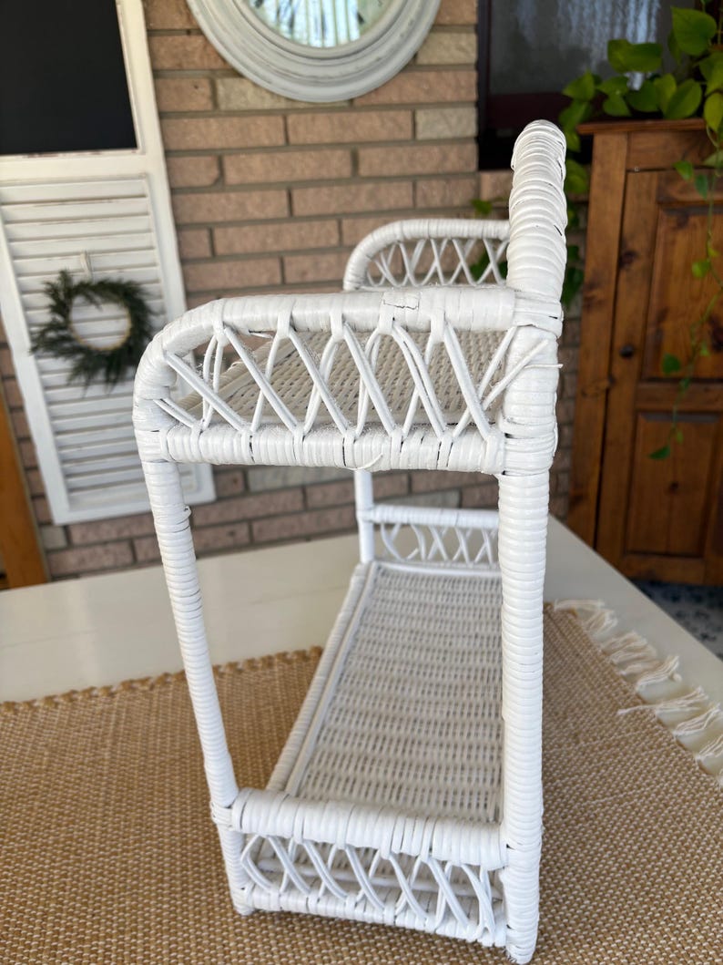 May include: A white wicker shelf with a rectangular base and a curved top. The shelf features a woven design and is supported by vertical posts. The piece is designed for storage or display and has a vintage aesthetic.