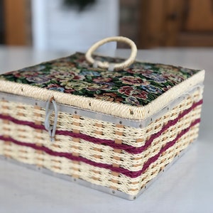 May include: A woven wicker storage box with a floral tapestry lid and a metal handle. The box has a burgundy and cream striped pattern.
