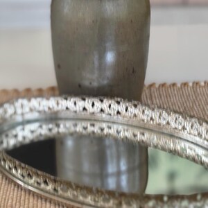 May include: An oval-shaped vanity tray with a reflective mirrored base and a raised, metallic silver-toned border featuring an intricate, repeating openwork pattern. The tray is positioned horizontally, showcasing its polished surface and detailed rim.
