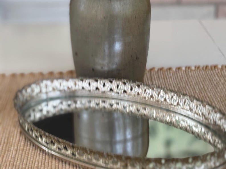 May include: An oval-shaped vanity tray with a reflective mirrored base and a raised, metallic silver-toned border featuring an intricate, repeating openwork pattern. The tray is positioned horizontally, showcasing its polished surface and detailed rim.