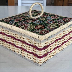 May include: A woven wicker storage box with a floral fabric lid and a metal ring handle. The box has a cream and burgundy color scheme.