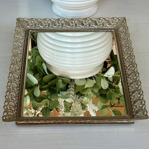 May include: A silver tray with a mirrored surface and an ornate, decorative frame. The tray is reflecting a white ceramic vase with a textured design.