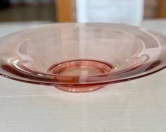 May include: A pink glass bowl with a wide, shallow base and a slightly raised center. The bowl has a smooth, glossy finish.