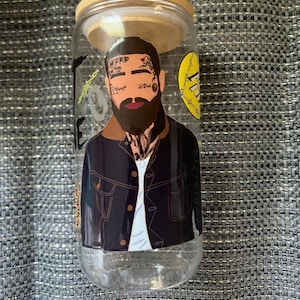 May include: A clear glass jar with a bamboo lid featuring a sticker of a man with tattoos and a beard. The man is wearing a black denim jacket and a white t-shirt. The sticker has a yellow circle with a barbed wire design and the text "Free".
