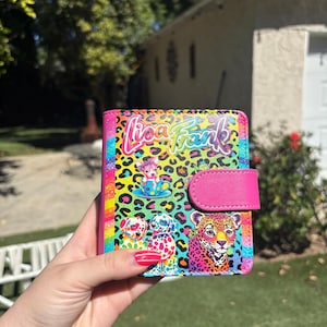 May include: A rectangular wallet featuring a vibrant Lisa Frank logo and a colorful leopard print background. The design includes illustrations of a puppy in a teacup, two spotted dogs, and a leopard, with a solid pink snap closure tab on the side.