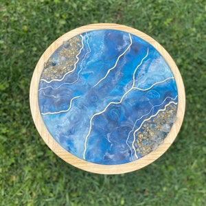 May include: Round wooden table with a blue resin top. The resin design features swirling shades of blue with gold accents and embedded crystals. The table has a natural wood edge.