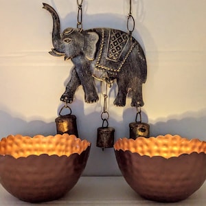 May include: A decorative hanging wind chime featuring an elephant design with intricate details. The chime has two copper-colored bowls at the bottom.