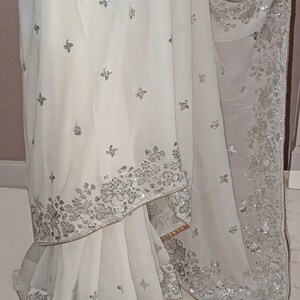 May include: A white chiffon saree with silver sequins and embroidery. The saree has a floral design and a scalloped border.