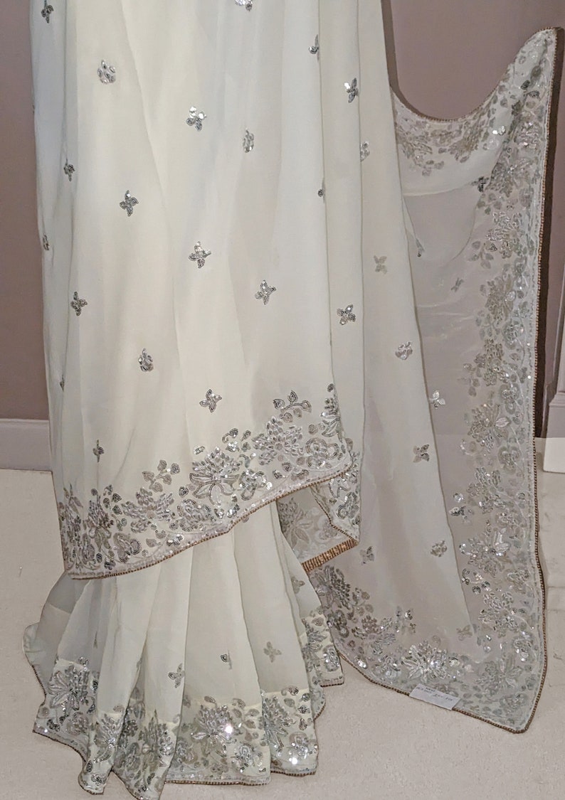 May include: A white chiffon saree with silver sequins and embroidery. The saree has a floral design and a scalloped border.