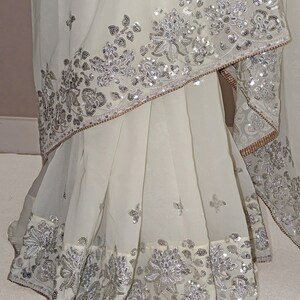 May include: A white chiffon saree with silver sequins and embroidery. The saree has a floral design and is decorated with a border of sequins.