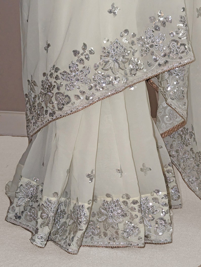 May include: A white chiffon saree with silver sequins and embroidery. The saree has a floral design and is decorated with a border of sequins.
