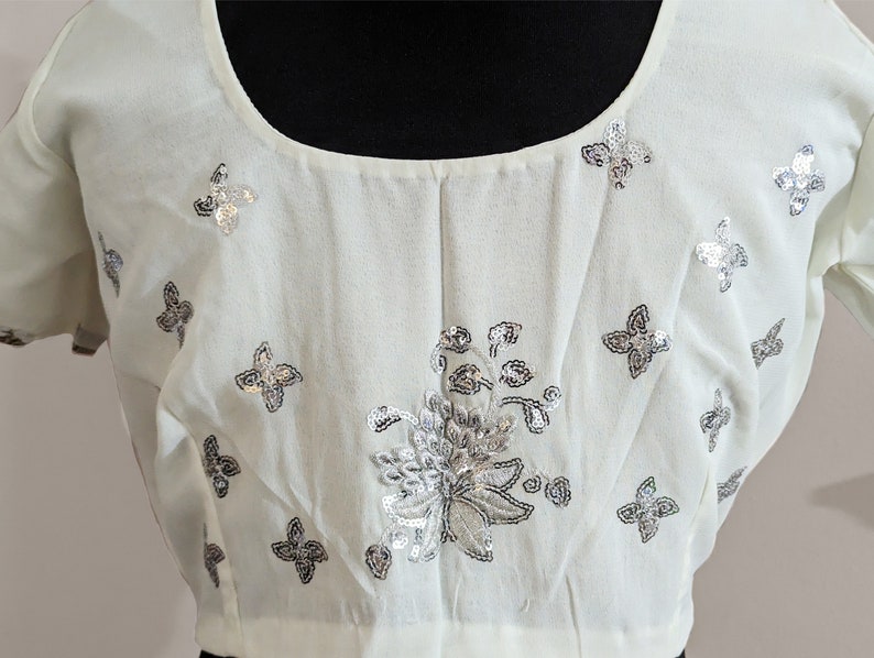 May include: A white blouse with a scoop neck and short sleeves. The blouse is decorated with silver sequins in a floral pattern and butterfly shapes.