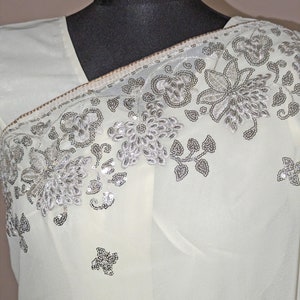 May include: A white sheer saree with silver sequins and floral embroidery. The saree is draped over a black mannequin.