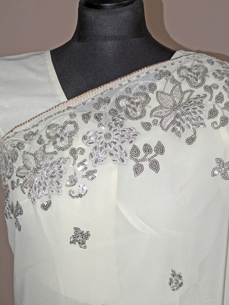 May include: A white sheer saree with silver sequins and floral embroidery. The saree is draped over a black mannequin.