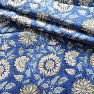 Può includere: Primo piano di tessuto blu con motivo floreale. Il design presenta grandi fiori bianchi e verde chiaro con foglie e steli dettagliati. Il tessuto è piegato, mostrando la consistenza e la profondità del materiale.