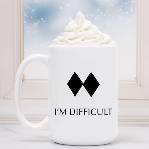 May include: White ceramic coffee mug with a black double diamond symbol and the text "I'M DIFFICULT". The mug is filled with a white whipped cream topping.