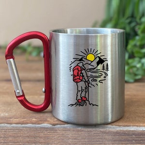 May include: A stainless steel travel mug with a red carabiner handle. The mug features a graphic of a hiker with a backpack standing on a mountaintop, with a sun and mountains in the background.