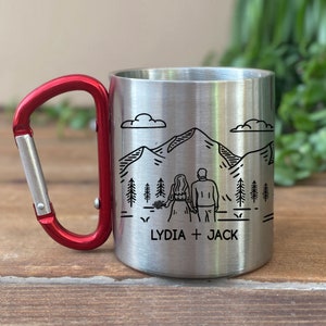 May include: A stainless steel travel mug with a red carabiner handle. The mug is engraved with a mountain scene and the text "LYDIA + JACK".