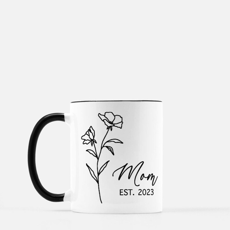 May include: White ceramic mug with a black rim and handle. The mug has a black line drawing of a flower with the text "Mom Est. 2023" on the front.