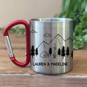 May include: A stainless steel camping mug with a red carabiner handle. The mug features a black and white illustration of mountains, trees, a tent, and a campfire. The text "LAUREN & MADELINE" is printed on the mug.