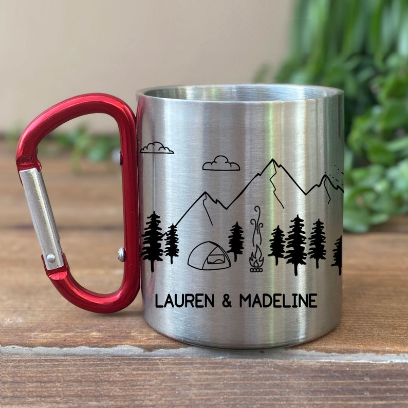 May include: A stainless steel camping mug with a red carabiner handle. The mug features a black and white illustration of mountains, trees, a tent, and a campfire. The text "LAUREN & MADELINE" is printed on the mug.