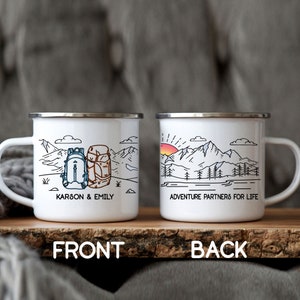 May include: Two white enamel mugs with black outlines of mountains, trees, and a lake. One mug has two backpacks and the text "KARSON & EMILY". The other mug has a sunrise and the text "ADVENTURE PARTNERS FOR LIFE".