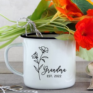 May include: White enamel mug with a black outline and a blue rim. The mug has a black line drawing of a flower with the text "Grandma Est. 2022" printed on it.
