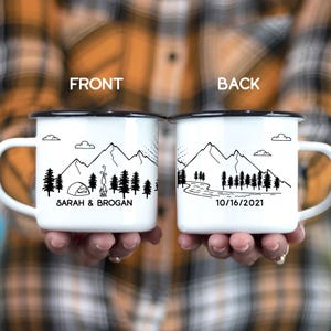 May include: Two white enamel mugs with black rims. The front mug features a mountain scene with a tent and campfire and the text "SARAH & BROGAN". The back mug features a mountain scene with a river and the date "10/16/2021".