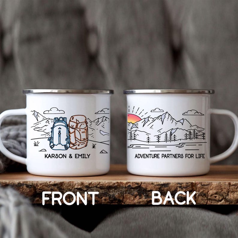 Puede incluir: Dos tazas de camping de esmalte blanco con bordes plateados. Una taza presenta una escena de monta&ntilde;a con dos mochilas y los nombres "Karson & Emily". La otra taza tiene un dise&ntilde;o de monta&ntilde;a y puesta de sol con el texto "Adventure Partners For Life."