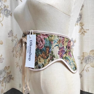 May include: A floral tapestry corset with a light pink trim and ribbon tie. The corset is displayed on a mannequin. The tag reads "Dikesshop on Etsy". The tapestry features a variety of colors including pink, green, blue, and yellow.