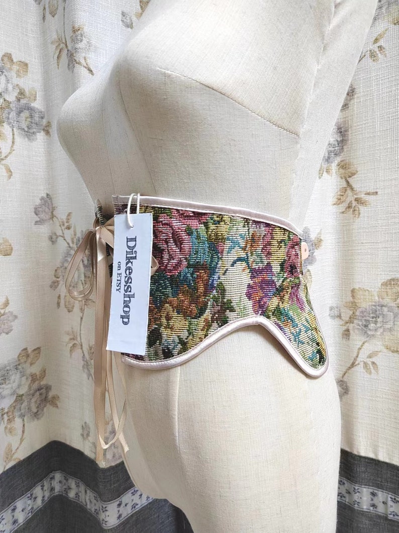 May include: A floral tapestry corset with a light pink trim and ribbon tie. The corset is displayed on a mannequin. The tag reads "Dikesshop on Etsy". The tapestry features a variety of colors including pink, green, blue, and yellow.