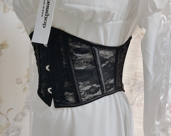 May include: Black lace corset with silver eyelets and a black satin trim. The corset is worn over a white blouse with ruffled sleeves. The tag on the corset reads "Dikessshop on Etsy".