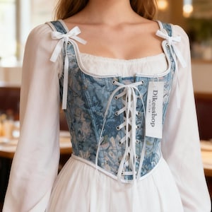 May include: A blue floral corset with white lacing and a white trim, paired with a white blouse. The corset has a square neckline and is adorned with white bows on the straps. The tag reads "Dikesshop".