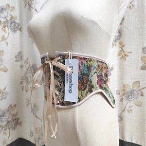 May include: A tapestry corset with a floral pattern in shades of green, red, and purple, displayed on a mannequin. The corset has a beige trim and a ribbon closure. A tag reads "Dxesshop on Etsy".