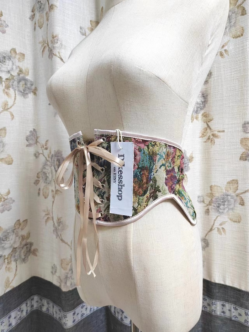 May include: A tapestry corset with a floral pattern in shades of green, red, and purple, displayed on a mannequin. The corset has a beige trim and a ribbon closure. A tag reads "Dxesshop on Etsy".