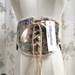 May include: A floral tapestry corset with a cream-colored trim and ribbon closure. The corset is displayed on a mannequin. A tag with the text "Dikesshop on Etsy" is attached.