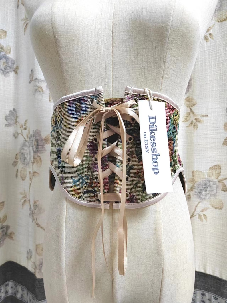 May include: A floral tapestry corset with a cream-colored trim and ribbon closure. The corset is displayed on a mannequin. A tag with the text "Dikesshop on Etsy" is attached.