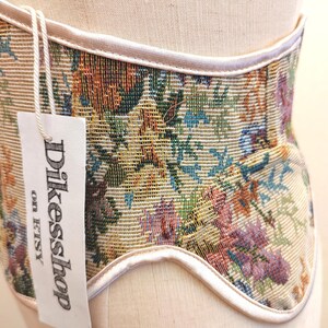 May include: A beige corset with a floral tapestry design in shades of green, orange, blue, and purple. The corset has a white trim and a tag that reads "Dikesshop on Etsy".