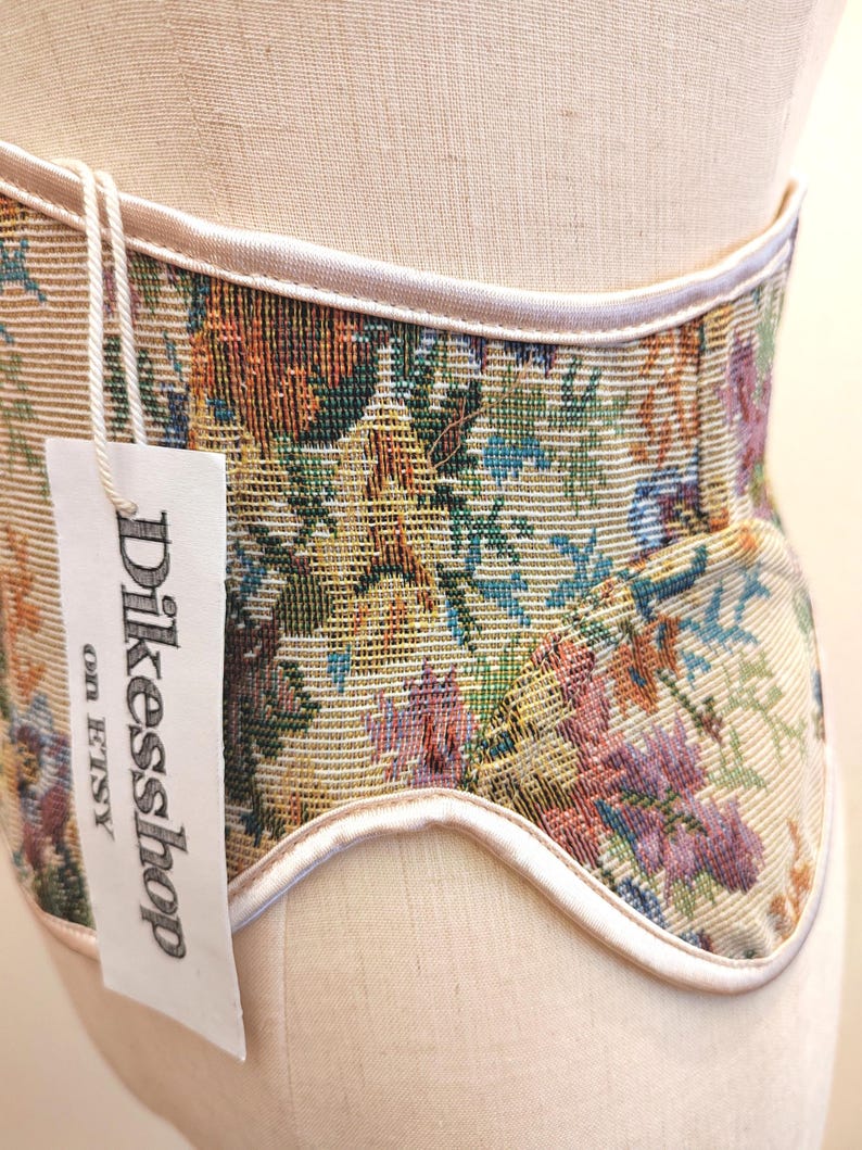 May include: A beige corset with a floral tapestry design in shades of green, orange, blue, and purple. The corset has a white trim and a tag that reads "Dikesshop on Etsy".