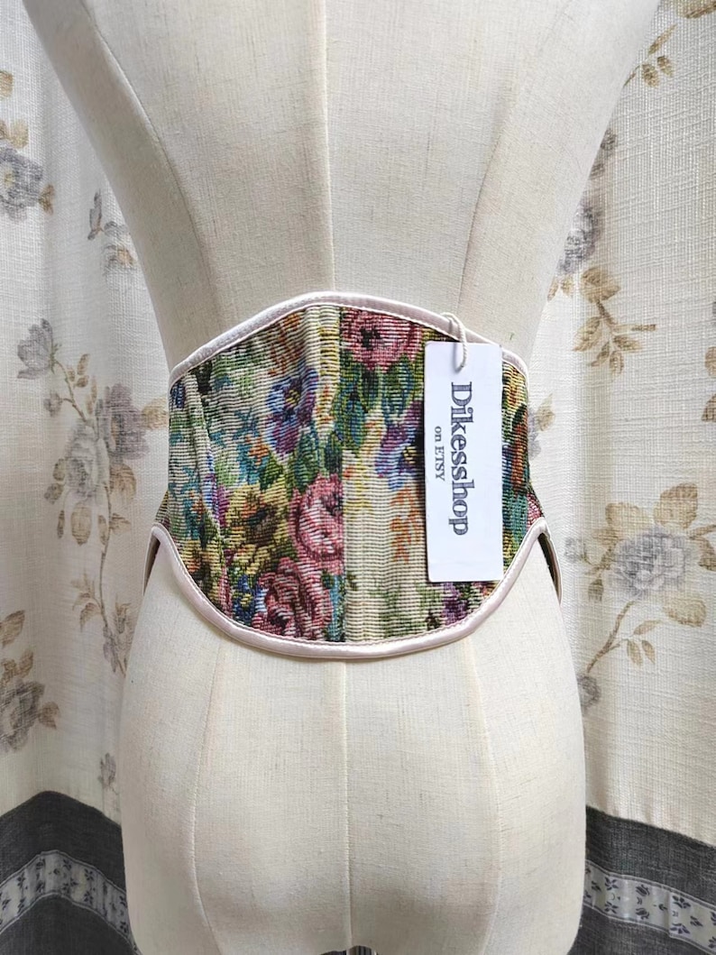 May include: A floral tapestry corset with a cream-colored trim and a tag that reads "Dikesshop on Etsy." The corset is displayed on a mannequin. The floral pattern includes pink, blue, and green hues.