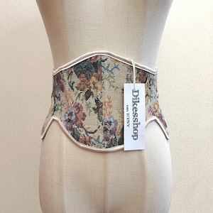 May include: A floral tapestry corset belt with a cream-colored trim. The belt features a variety of flowers in shades of red, purple, green, and yellow. A tag with the text "Dikesshop on Etsy" hangs from the belt.