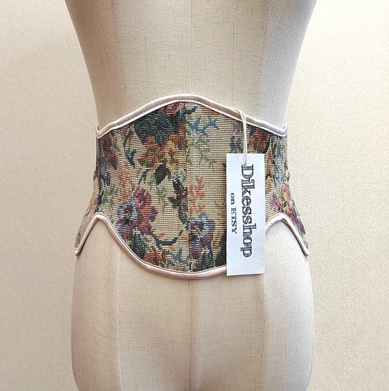 May include: A floral tapestry corset belt with a cream-colored trim. The belt features a variety of flowers in shades of red, purple, green, and yellow. A tag with the text "Dikesshop on Etsy" hangs from the belt.