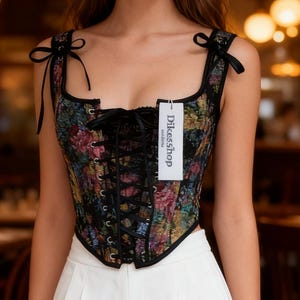 Könnte beinhalten: Ein floral bedrucktes Korsett mit schwarzem Besatz und Schnürung vorne. Das Korsett hat schwarze Träger, die an den Schultern gebunden werden. Ein Etikett mit der Aufschrift "Dikesshop" ist am Korsett befestigt.