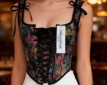 Könnte beinhalten: Ein floral bedrucktes Korsett mit schwarzem Besatz und Schnürung vorne. Das Korsett hat schwarze Träger, die an den Schultern gebunden werden. Ein Etikett mit der Aufschrift "Dikesshop" ist am Korsett befestigt.