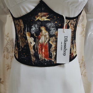 May include: A black corset with a colorful Renaissance-inspired print featuring a woman and a cherub. The corset has a black elastic band at the top and bottom. A white tag with the text "Dikesshop on Etsy" is attached to the corset.