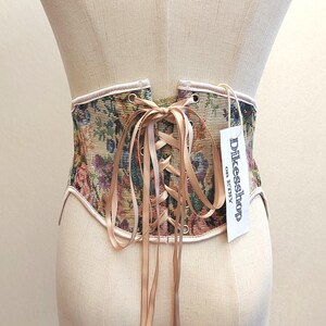May include: A tapestry corset with a floral pattern in shades of green, purple, and yellow. The corset has a beige satin ribbon for lacing and a tag that reads "Dikesshop on Etsy". The edges are trimmed with a light pink fabric.
