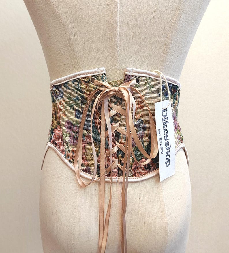 May include: A tapestry corset with a floral pattern in shades of green, purple, and yellow. The corset has a beige satin ribbon for lacing and a tag that reads "Dikesshop on Etsy". The edges are trimmed with a light pink fabric.